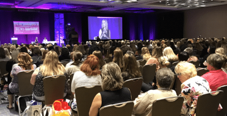 View from behind an audience sitting in a conference room watching a woman speak on stage